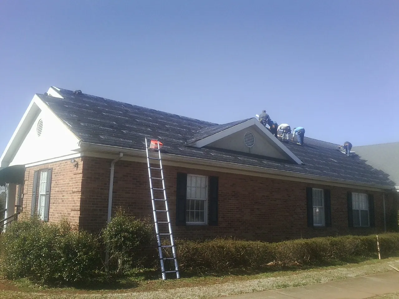 Crew tearing shingles off a brick ranch roof with a ladder set — Emergency Roof Repair work by our crew in Center Harbor