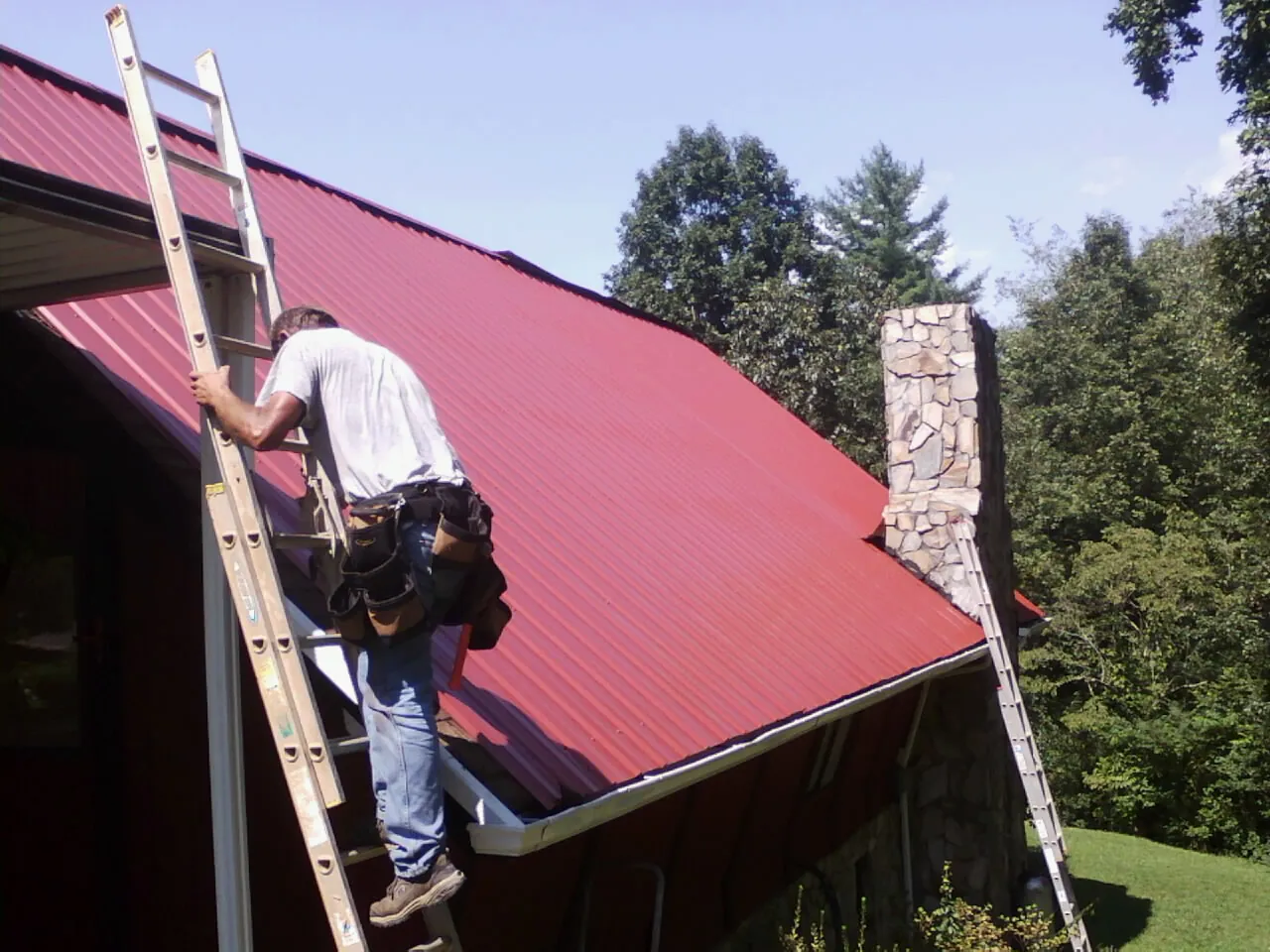 Roofer on a ladder beside a red metal roof and stone chimney — Soffit & Fascia Repair by Roofing Brothers in Center Harbor